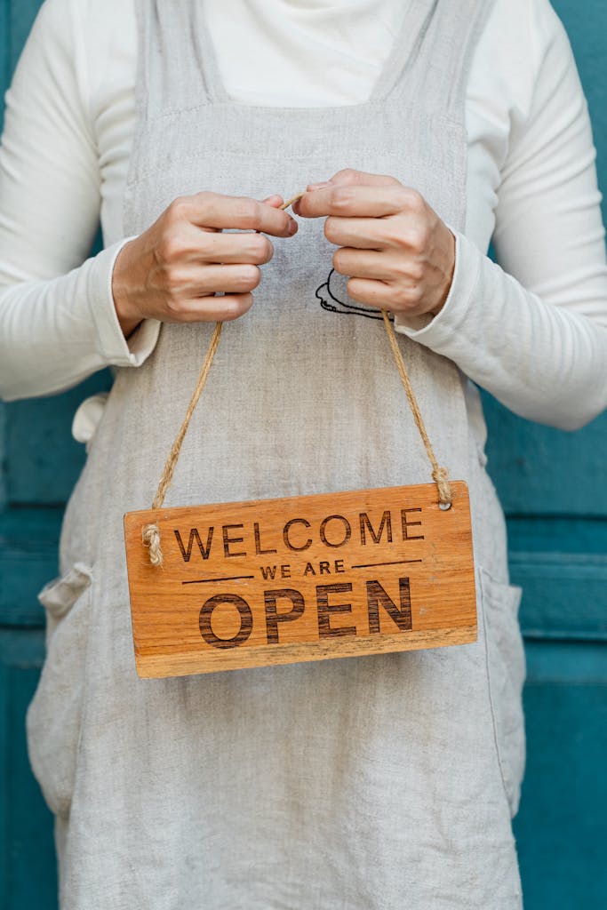 Crop faceless female in casual clothes and apron demonstrating wooden sign with text welcome we are open against blue door